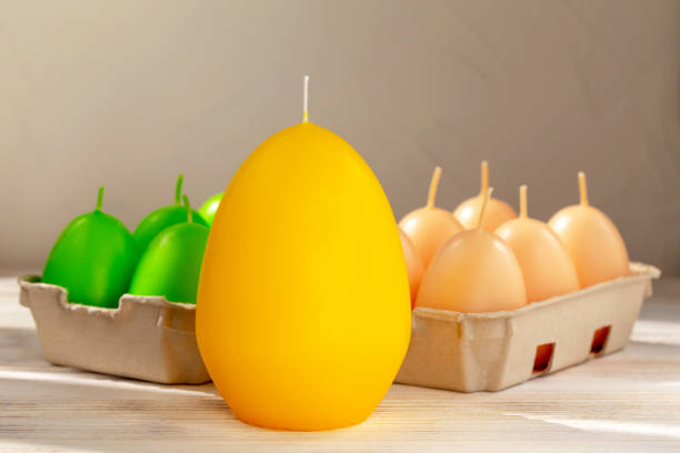 Easter. A large yellow egg-shaped candle stands on the table. Behind it are green and beige egg-shaped candles in trays. Close-up