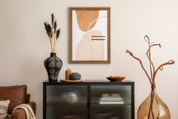 istock Creative composition of living room interior with mock up poster frame, black commode, vase with dried flowers, brown sofa, plaid and stylish personal accessories. Home decor. Template. 1457704343