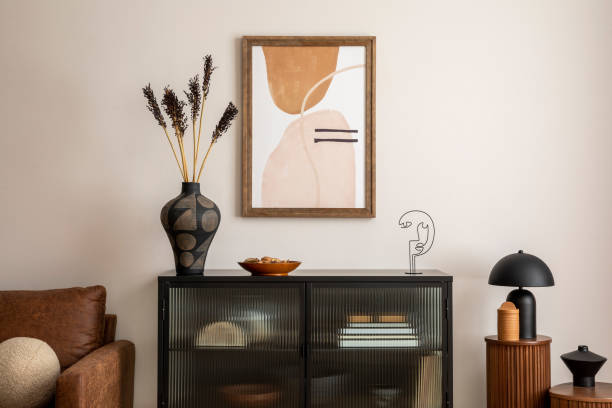 istock Creative composition of living room interior with mock up poster frame, black commode, vase with dried flowers, lamp on the wooden stand, brown sofa and personal accessories. Home decor. Template. 1457704386