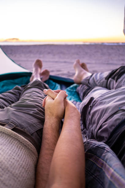 para trzymająca się za ręce obserwuje wschód słońca na cichej plaży z wnętrza namiotu. punkt widzenia. - sunrise beach couple hiking zdjęcia i obrazy z banku zdjęć