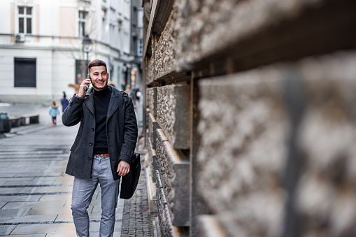 A successful and confident younger man is walking through the city and talking using his smartphone.