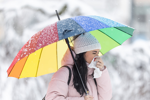 Woman with an umbrella outdoors sneezes into a napkin, getting cold from the bad winter weather.