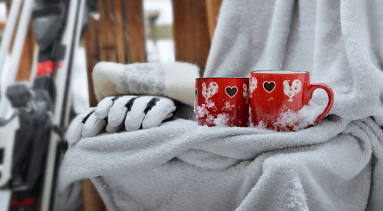 two red mugs with heart shaped on a plaid put on a chair  in  snow and ski equipment on terrace