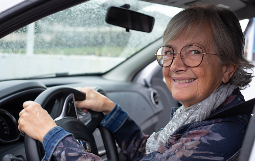Senior woman driver driving her car on a rainy day. Elderly woman holding the steering wheel looking smiling at camera