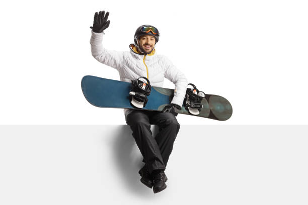 Man in a white jacket with a snowboard waving and sitting on a blank panel isolated on white background