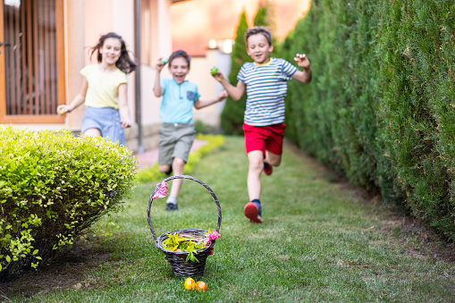 Three children are excitedly running in a back yard towards a pretty decorated basked filled with chocolate eggs for Easter
