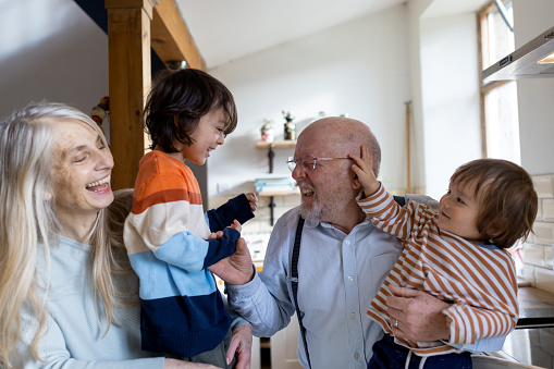 A waist-up shot of a senior married couple with their two grandchildren. They are laughing together standing in the kitchen.