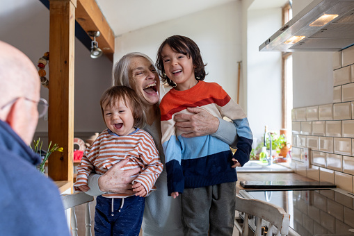 A waist-up shot of a grandmother with her arms around her two grandchildren. She is laughing with positive emotion. They are looking at their grandad.