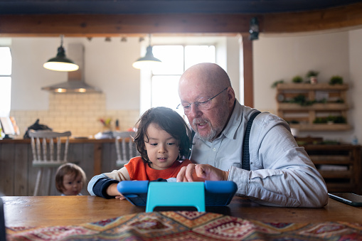 A full shot of a grandfather and grandson both playing and looking at a digital tablet sitting down at the kitchen table. The grandad has his arm around his grandson typing on the digital tablet with his other hand.