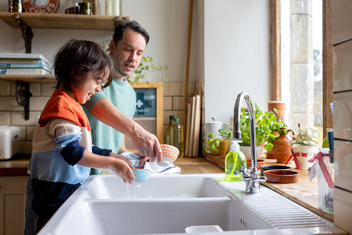 A waist-up shot of a male child and his dad washing and drying dishes together in their home. They are both smiling and wearing casual clothing.