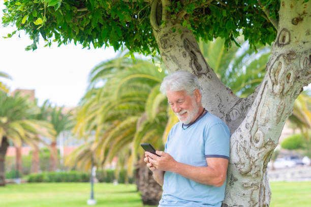 istock Beautiful bearded senior man standing under a tree in public park using mobile phone. Smiling elderly man holding telephone using app and social media 1457722504
