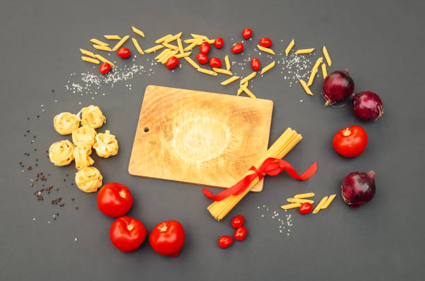 Cutting board surrounded by fresh tomatoes and onions, raw spaghetti, penne and fettuccine and seasonings on gray background. Top view, copy space.