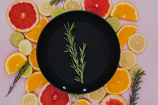 black frying pan with a sprig of rosemary in it on top of sliced citrus herbs. Oranges, grapefruit, lemon, lime on a pink background. flat lay