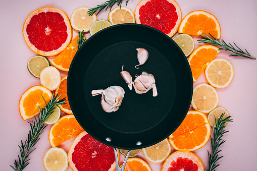 Black frying pan with garlic in it on sliced citrus herbs. Oranges, grapefruit, lemon, lime on a pink background. flat lay