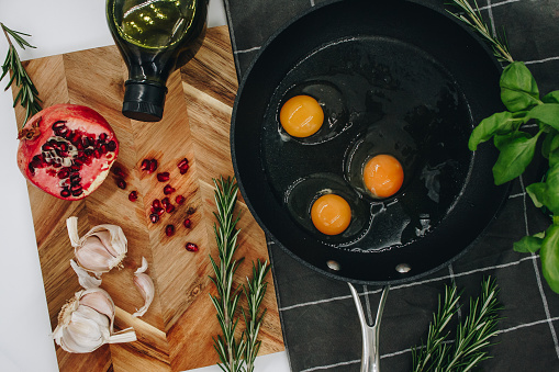 black frying pan on a black towel with three raw eggs in it next to a wooden cutting board with a red pomegranate on it.