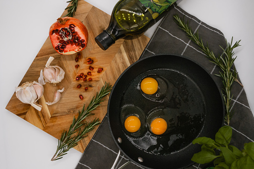 black frying pan on a black towel with three raw eggs in it next to a wooden cutting board with a red pomegranate on it.