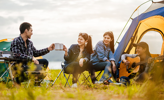 Team of asian climbers hiker are sitting and enjoying a drink after a set up outdoor tent playing music together in the forest path autumn season.hiker, team, forest, camping , activity concept.