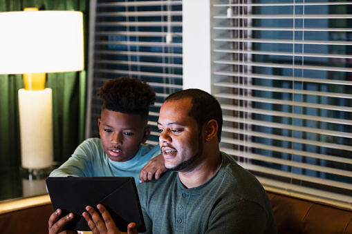 A multiracial man and his stepson sitting together at night on the living room couch, watching something on a digital tablet. The boy is 10 years old, African-American and Native American. His stepfather is African-American, Native American, and Hispanic. The main focus is on dad.
