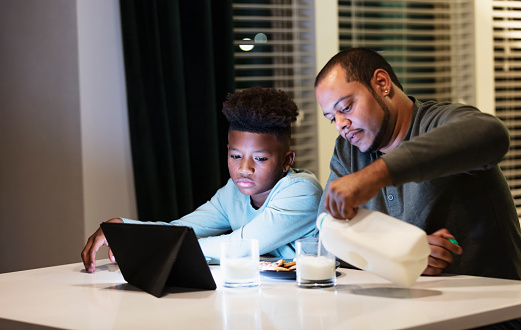 A multiracial boy and his stepfather sitting together at the kitchen counter watching TV or a movie on a digital tablet at night. They are having milk and cookies. The boy is 10 years old, African-American and Native American. His stepfather is African-American, Native American, and Hispanic. The main focus is on the boy.