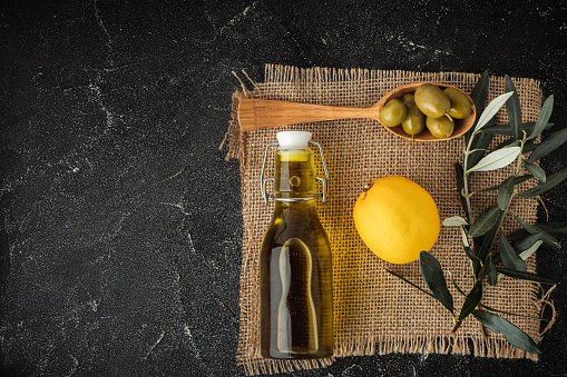 Green olives in a wooden spoon next to a bottle with olive oil and leaves on sackcloth. Black background. \nBottle of cold pressed oil. Traditional Greek and Italian food. Flat lay. Copy space
