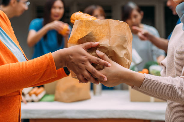 Unrecognizable woman hands out food donations during charity drive An unrecognizable woman hands a bag of donated food to someone during a community food drive. An Asian female volunteer and her colleagues distributing grocery food at community food bank food bank stock pictures, royalty-free photos & images