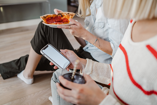 Close-up shot of female friends enjoying pizza at home. One of them is eating the other one is holding smart phone with a white screen.