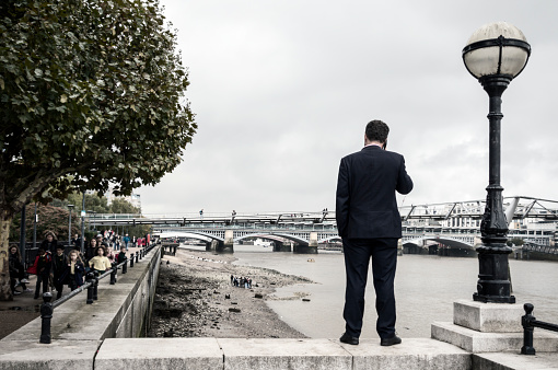 A businessman talking on his mobile phone whilst standing on the edge of a flight of steps leading down to the edge of the River Thames at Bankside, London. (Subdued colour.)