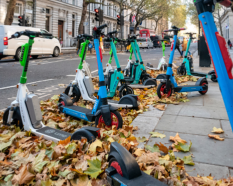 An assortment of rental e-scooters parked amongst the autumn leaves by the side of Northumberland Avenue in the City of Westminster, London.
