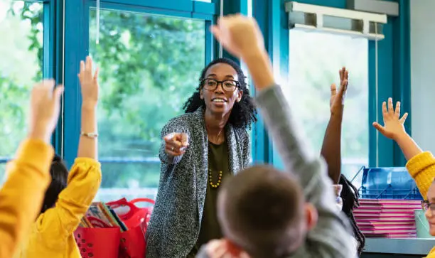 A multiracial group of elementary students in class raising their hands to answer a question. The focus is on their teacher, an African-American and Hispanic woman in her 30s, who is standing in front, facing them, smiling and pointing to a student. Most of the students are 9 years old.