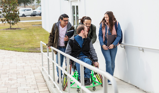 A multiracial group of five college students walking to class together, using a wheelchair ramp to get into a building since one of them is in a wheelchair. He has cerebral palsy.