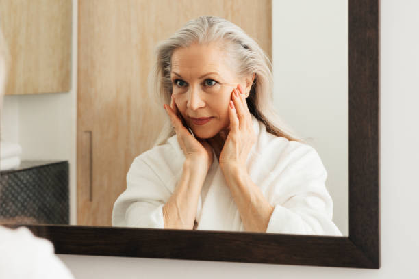 istock Senior woman in a bathrobe looking at a mirror and touching the face 1457744442
