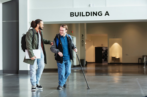 Two multiracial university students walking in a school building, conversing. The young men are in their 20s. The one on the right using a walking cane has cerebral palsy.