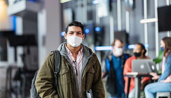A university student looking at the camera, standing in the foreground, with his friends studying in the background out of focus. The young Hispanic man, in his 20s, is wearing a protective face mask trying to prevent the spread of coronavirus during the covid-19 pandemic.