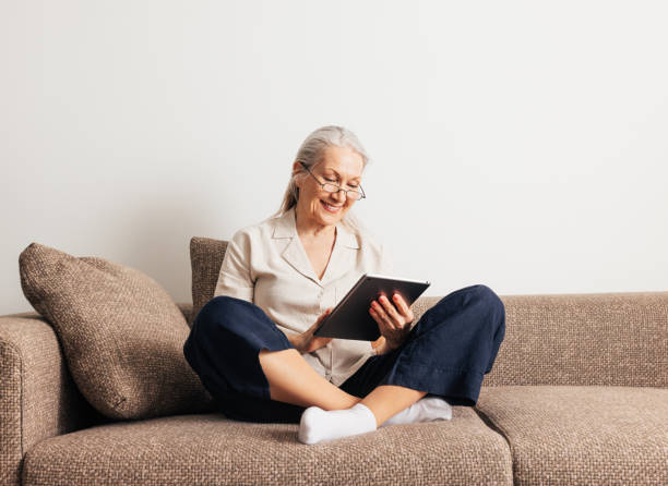 istock Cheerful aged female in glasses relaxing on a couch holds a digital tablet 1457747005