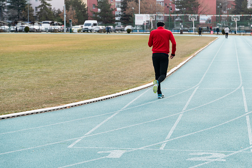 young man doing sports on the running track. young man wearing red and black tracksuit running on public sports ground. 
Taken with a full-frame camera in daylight