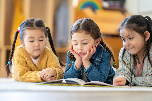 Three female kindergarten students lay on the floor, side-by-side, as they hold themselves up on their elbows and read a book together.  They are each dressed casually and paying close attention to the words.