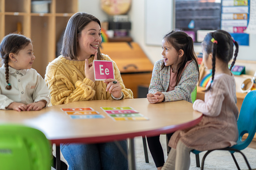 A Kindergarten teacher sits with a small group of students as she teaches them the different sounds each letter of the alphabet makes.  Each of the students are dressed casually and paying close attention to their teacher.