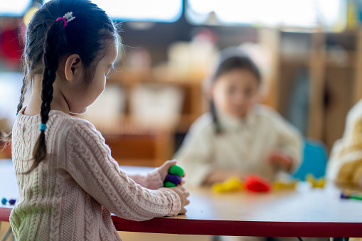 A sweet little girl of Asian decent sits at a table in her Kindergarten classroom as she plays with Playdoh.  She is dressed casually and is surrounded by her classmates who are also creating with Playdoh.