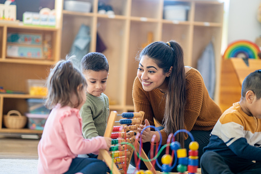 A female Kindergarten teacher of Middle Eastern decent, sits on the floor with students as they play with various toys and engage in different activities.  They are each dressed casually as they learn through their play.