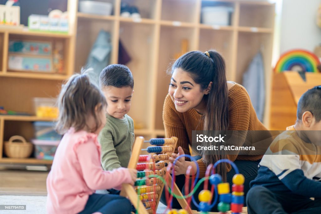 Learning Through Play A female Kindergarten teacher of Middle Eastern decent, sits on the floor with students as they play with various toys and engage in different activities.  They are each dressed casually as they learn through their play. Teacher Stock Photo