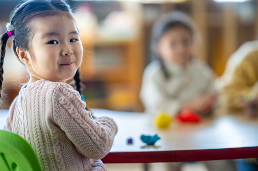 A sweet little girl of Asian decent sits at a table in her Kindergarten classroom as she plays with Playdoh.  She is dressed casually and is surrounded by her classmates who are also creating with Playdoh.