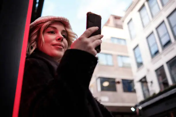 A young woman is standing near neon light and she is using smart-phone. She is smiling and wearing winter clothes. Technology addition concept.