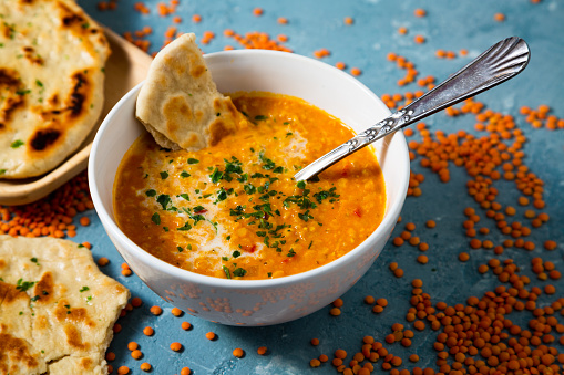 Red lentil soup with coconut milk and curry accompanied by naan bread sorrounded by uncooked lentils on a blue background
