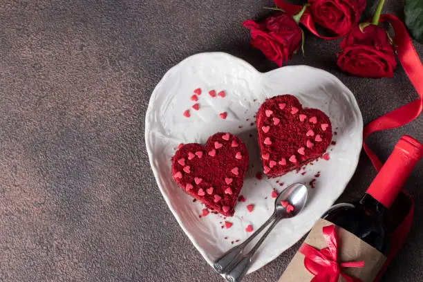 Photo of Cakes Red velvet in the shape of hearts on white plate, roses and bottle wine for Valentines Day on brown background, Top view