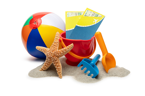 This is a close up photo of a children's colorful striped beachball next to a plastic toy bucket shovel and rake sitting on sand and photographed in the studio isolated on a white background.