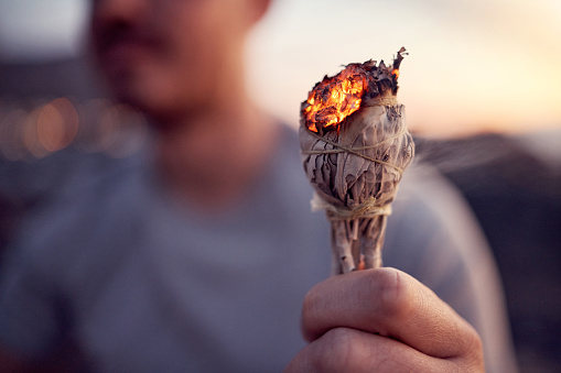 Man, hand and burning sage for a spiritual meditation on the beach at sunset for health and wellness. Smudge, burn herb and alternative healing ceremony to cleanse chakra in a meditating cleansing