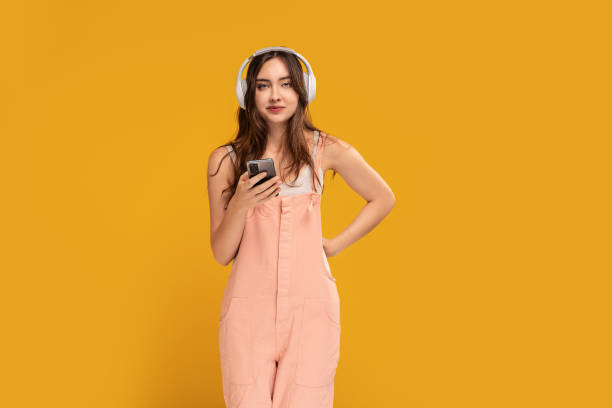 istock Female student using smartphone and headphones, looking at the camera. Studio shot, yellow background, Happy young woman. Online app in mobile phone. 1457778283