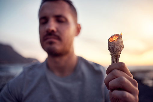 Man, hand and burning sage on the beach while meditating for spiritual peace and zen mindfulness. Herb, burn and alternative cleanse ceremony to meditate for smudge during a seaside peaceful time