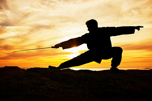 Sunset, sport and man doing tai chi with a sword on a hill for exercise and fitness workout. Silhouette, training and sports exercising male at sunrise for chi gong practice for health and wellness