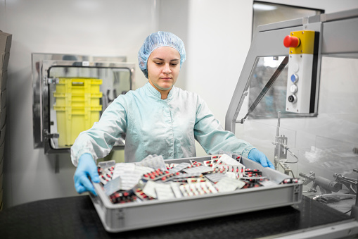 A young female worker in protective gear works diligently on a pharmaceutical production line, packaging capsules into blister packs. A look into the hard work and attention to detail that goes into producing life-saving medications, even when working alone.